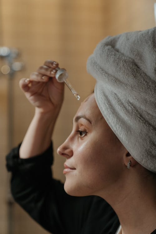 Profile image of a woman with a towel wrapped around her head holding a dropper over her forehead, depicting a night time skincare routine.