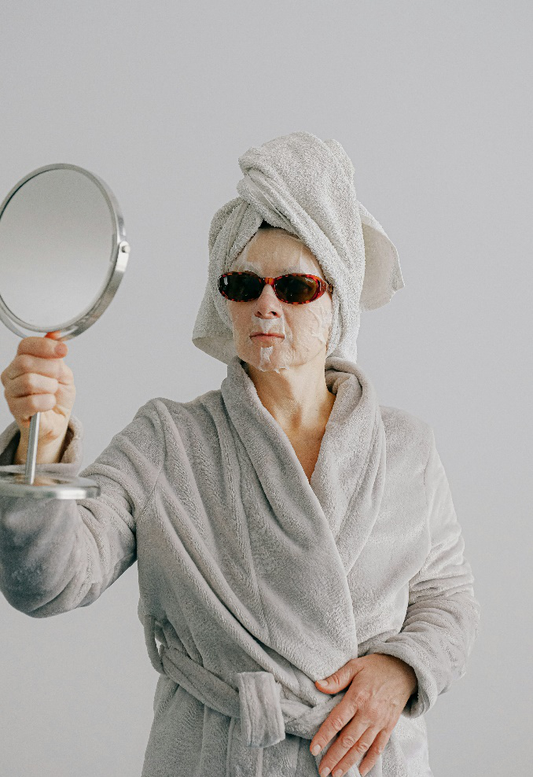 Image of a woman wearing a bathrobe, a face mask, and sunglasses, looking at a handheld mirror.